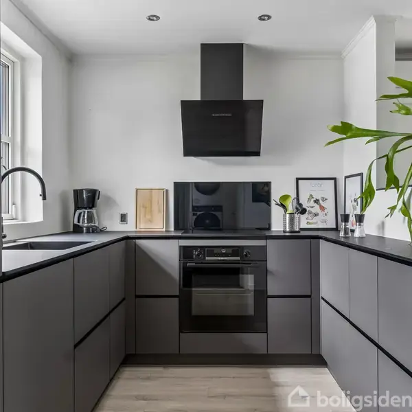Et moderne køkken med grå skabe er i fokus, udstyret med en ovn. En grøn plante dekorerer rummet, og en kaffemaskine står ved vasken nær et vindue.