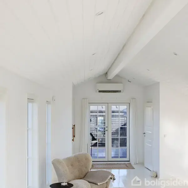 Klimaanlæg hænger på væggen over en dobbelt fransk dør. Rummet har hvidmalede vægge og loft med synlige bjælker. Udenfor ses en terrasse med stole og et bord.