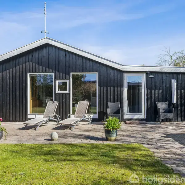 Sort træhus med store vinduer, to liggestole foran på en flisebelagt terrasse. Omgivet af grøn græsplæne og buske under en blå himmel.