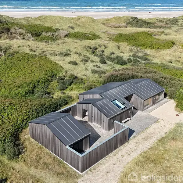 Et moderne sommerhus med sort facade står stille i grønne klitlandskaber. Baggrunden afslører en sandstrand og det blå hav i horisonten.
