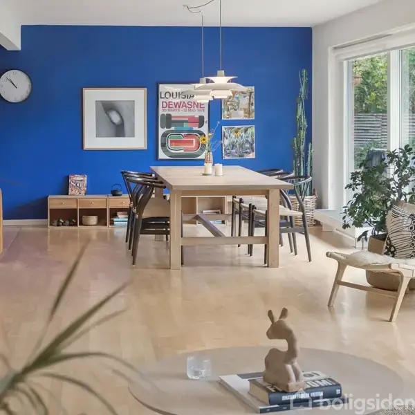 Et træbord med sorte stole står i en stue. Blå væg med malerier, et ur og reol. Store vinduer og flere planter skaber en lys, moderne atmosfære. Tekst: “LOUISE DEWASNE.”