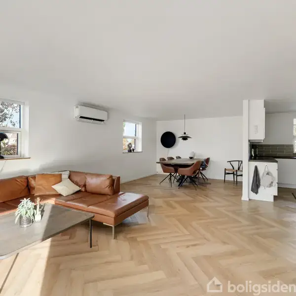 Brun lædersofa står i en stue med trægulv. Ved siden af er et glasbord, og i baggrunden ses et spisebord med stole og et køkken. Væggene er hvide.