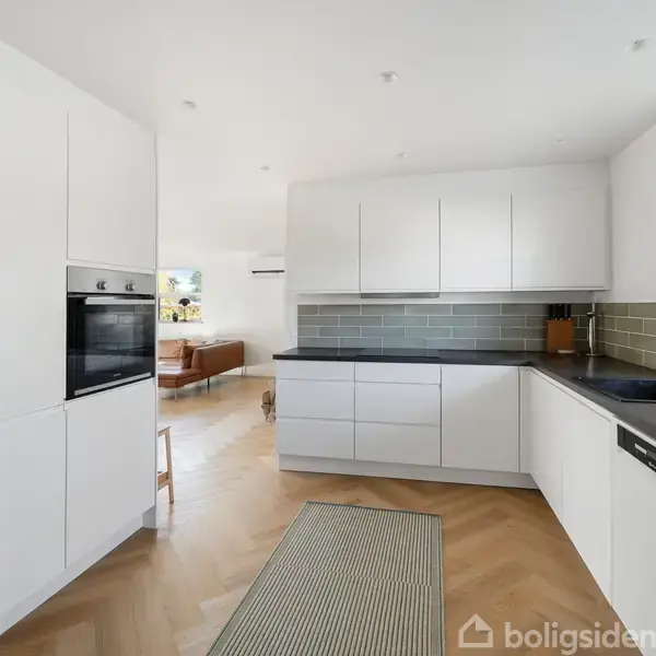 Køkken med hvide skabe og en sort bordplade, indbygget ovn, grønne fliser på væggen, og en stor vask. Det er i et lyst rum med trægulv og udsigt til en stue.