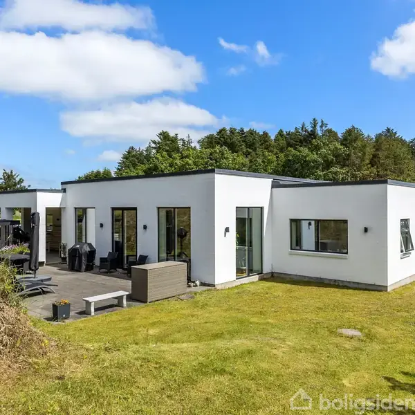 Hvid, moderne etplansvilla står på en grøn græsplæne, omgivet af træer og blå himmel. Store vinduer kendetegner designet. Terrasse med havemøbler ses i forgrunden.