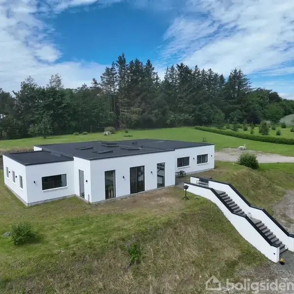 En moderne, hvid bungalow står på en grøn græsplæne. En ydre trappe fører op til huset. I baggrunden ses træer og en åben, blå himmel.