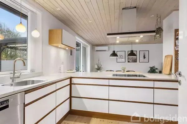 Moderne køkken med hvide skabe, stor køkkenø og indbygget ovn. To lamper hænger over spisebordet i stuen. Trægulve og træloft skaber en varm atmosfære. Billeder på væggen.