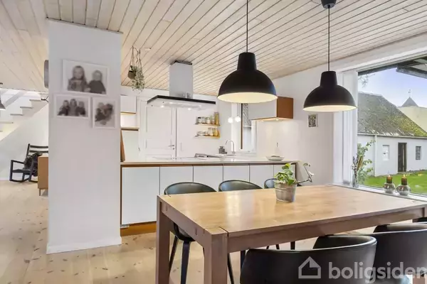 Spisebord og stole står i en lys spisestue med hvide vægge og træloft. To sorte lamper hænger over bordet. Et åbent køkken er i baggrunden, og der er billeder på væggen.