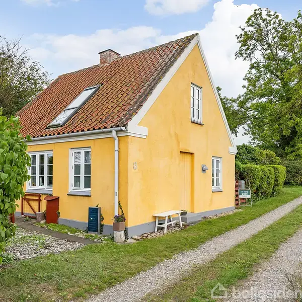 Et gult hus med rødt tag står ved en grusvej omgivet af grønne buske og træer; huset har hvide vinduer, og der er en lille bænk ved siden af hoveddøren.