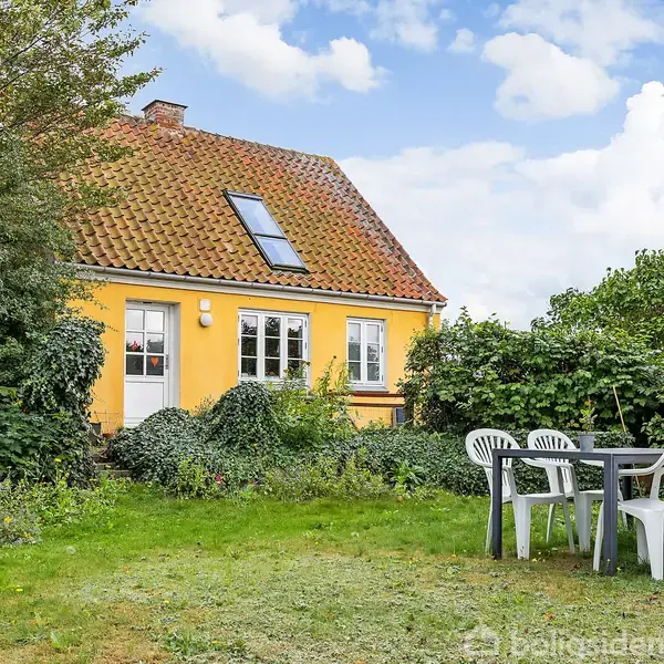 Et gulmalet hus med rødt tegltag har en dør og et skråt vindue. I haven foran står et bord omgivet af fem hvide plasticstole, omgivet af grønne buske og træer.