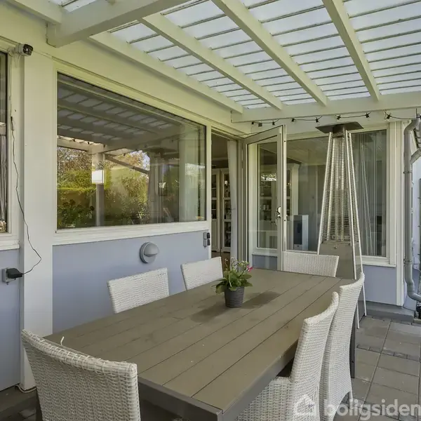 Udendørs spisearrangement består af et træbord med seks hvide kurvestole. Der er en plante i midten af bordet. Omkring er der en overdækket terrasse og glasvægge med udsigt til en have.