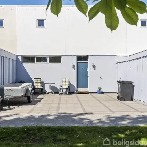 Et rækkehus med en blå dør står i en gårdsplads med fliser. Der er to stole og et bord, en trailer og en affaldsbeholder i gårdspladsen.
