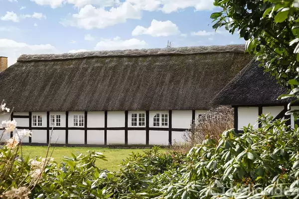 Bindingsværkshus med stråtag står i en have omgivet af buske. Huset har hvide vægge og adskillige små vinduer. Sollys lyser op, og skyer ses på himlen.