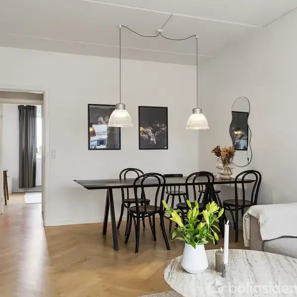 En stue med et sort spisebord, omgivet af sorte stole. To pendellamper hænger over bordet. Stuen er stilfuld med billeder og en buet spejl på væggen, samt en grå sofa.