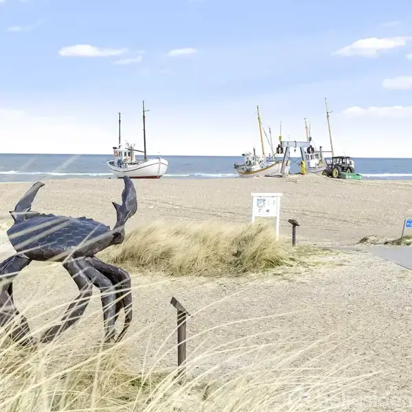 En metalskulptur af en krabbe står stationær på en sandstrand, mens flere fiskerbåde er trukket op på kysten i baggrunden. Skilte og vej ses i nærheden.