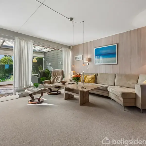 En stue med en beige hjørnesofa og et træbord placeret på et tæppe. Sofaen vender mod en stor glasdør med udsigt til have. En gulvlampe og billeder hænger på væggen.