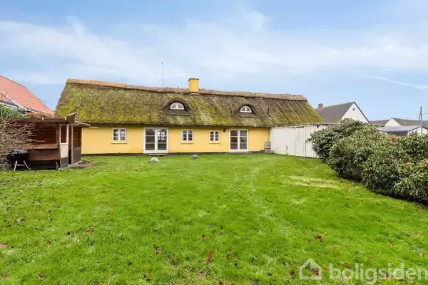 Et gult bindingsværkshus med stråtag står omgivet af en grøn have. Husets vinduer er hvide, og der er buske på højre side. I baggrunden ses flere huse.