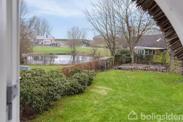 Grønt græs med buske omkranser en havedam, mens træer uden blade står langs hækken. I baggrunden ses et hus ved en landevej.