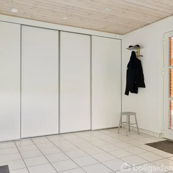 Et lyst rum med hvide skydedøre, flisegulv og træloft. En frakke og jakke hænger på knager, mens taburetter står ved siden af. En glasdør fører til en veranda.