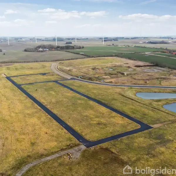Stor, rektangulær asfaltvej løber gennem en mark med gyldent græs. Omgivet af mindre søer og vindmøller i baggrunden under en klar himmel.
