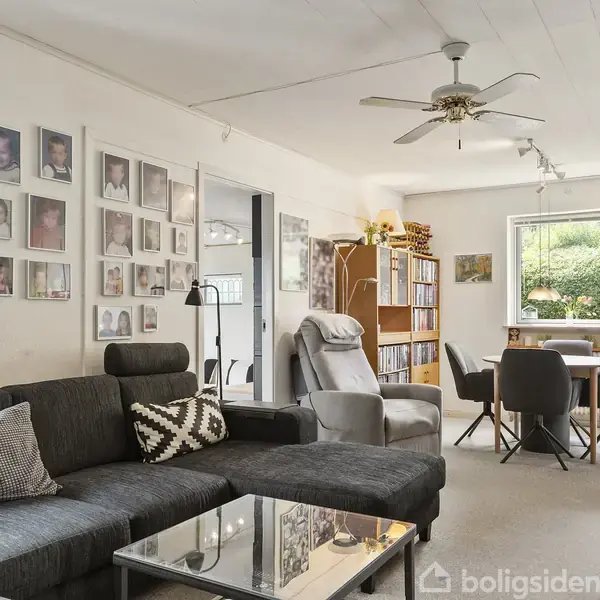 Stue med sort sofa, sofabord og lænestol. Fotovæg til venstre. Spisebord med stole foran vindue. Reol med bøger i baggrunden. Loftventilator hænger i midten.