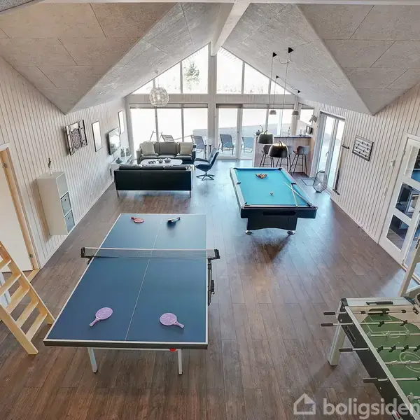 Bordtennisbord med net og pagajer i aktivitetstue med trægulv. I baggrunden ses et billardbord, et fodboldbord og sofaer med store vinduer og skrå lofter.