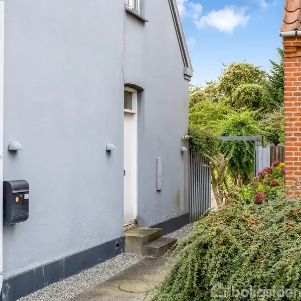 Grå husvæg ved siden af en rød murstensvæg. En smal sti med småsten fører mod en have, hvor grønne buske og et hegn er synlige. Skyfri himmel.