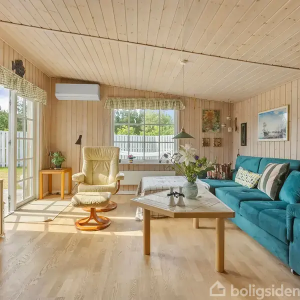 Stue med blå sofa og lænestol, sofabord med blomster, hvide trævægge, store vinduer, og udsigt til have gennem glasdør. Malerier og hylder dekorerer rummet.