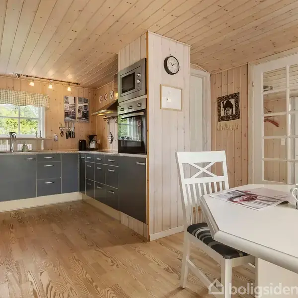 Et moderne køkken med grå skabe og indbyggede apparater. Et hvidt spisebord med stole står til højre. Træpaneler pryder væggene og loftet. En dør med glasruder fører til et andet rum.