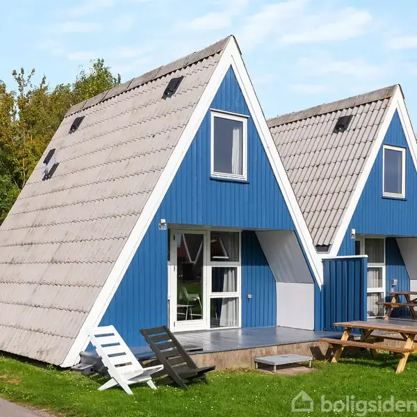 Blå A-formet hytte står stille på en græsbelagt grund ved siden af en sti. Et picnicbord og to hvide stole er placeret foran hytten.