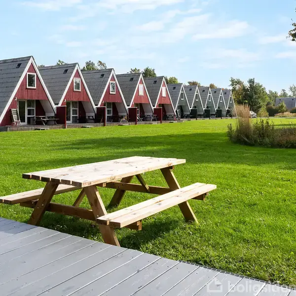 En række A-formede huse står i en linje på en græsplæne. Et træ picnicbord er placeret på en terrasse i forgrunden.