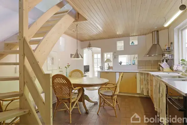 Et træspisebord med flettede stole står i et åbent køkken-/spiseområde. Til venstre fører en trætrappe op til en anden etage. Køkkenet har hvide elementer og stålemhætte.