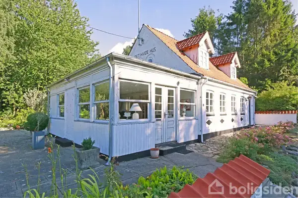 En hvidmalet villa med orangeri og rødt tegltag står blandt træer og buske. Teksten "DET HVIDE HUS" ses på facaden. Omkring huset er der en stenbelagt terrasse med blomsterbede.