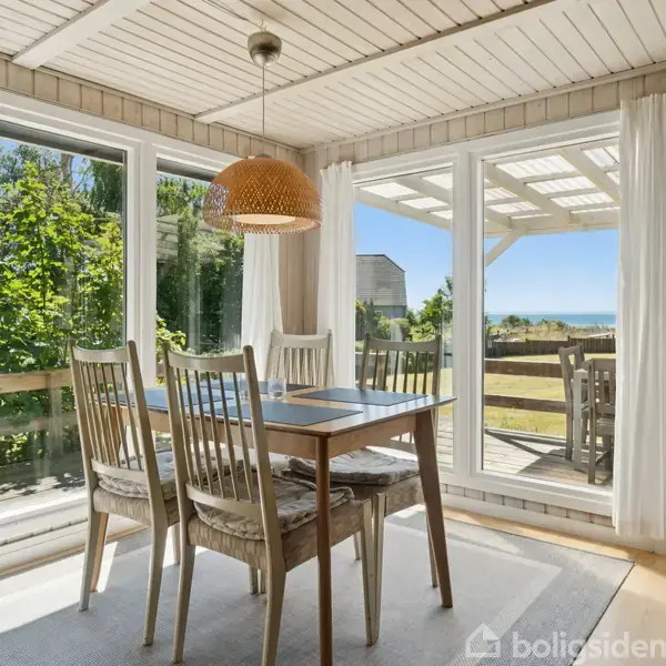 Et spisebord med stole står i en lys udestue med store vinduer. Sollys strømmer ind, udsigt til en have og havet i baggrunden. En hængelampe over bordet.