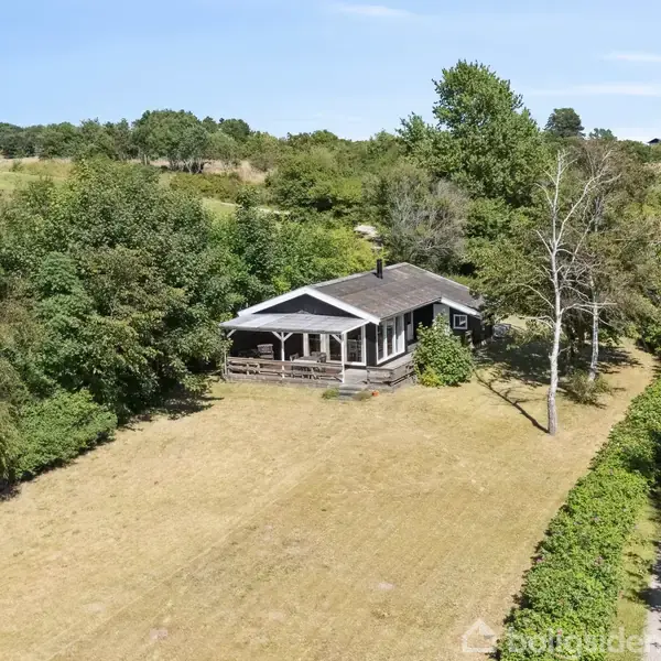 Et træhus står stille omgivet af grønne træer og græs i en åben landskabsscene. En smal sti løber langs højre side af billedet.