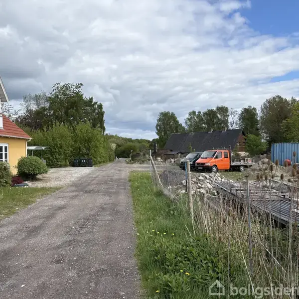Et gult hus står stille ved en grusvej. Ved siden af er der byggeudstyr og en orange varevogn. Omgivelserne er grønne med træer og buske under en skyet himmel.