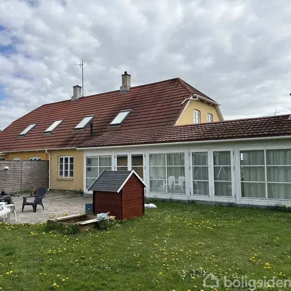 Et gult hus med rødt tegltag og flere vinduer, herunder en stor glaskarnap. I haven står nogle stole og et lille legehus på græsset under en delvist skyet himmel.