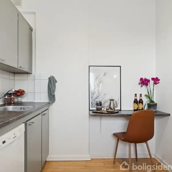 Køkken med grå skabe og opvaskemaskine. En bordplade til højre med en stol, blomster og drikkevarer står ved en hvid væg. Kogegrej ses også. Trægulv i rummet.