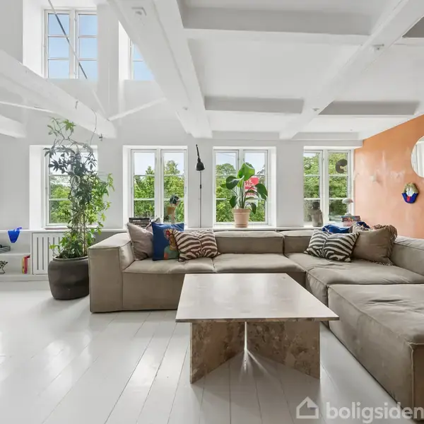 Stor, lys stue med en stor, grå hjørnesofa placeret foran flere høje vinduer. Et sofabord står i midten. Orange væg med spejl og kunst. Grønne planter i hjørnerne.