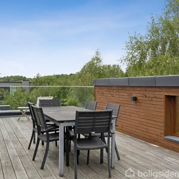 Et sort spisebord med seks stole står på en træterrasse. Ved siden af er liggestole. Baggrunden domineres af grønne træer mod en blå himmel.