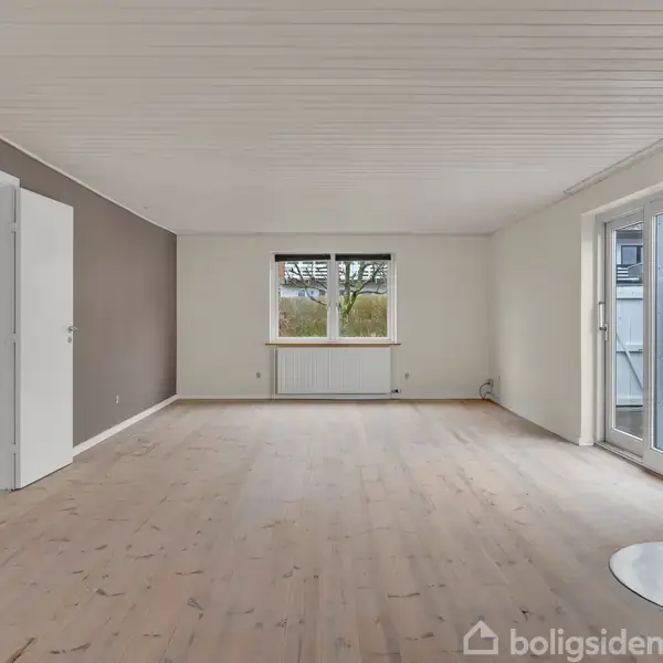 Stue med en brændeovn til højre, åben dør til venstre og vindue i baggrunden. Rum med lyse trægulve og hvide vægge.