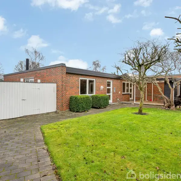 Rødt murstenshus med fladt tag står i en velplejet have med græsplæne og træer. En belagt sti fører til en hvid port. Skyfri himmel i baggrunden. Tekst: "boligsiden".