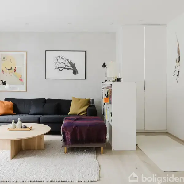 En stue med en mørk sofa dekoreret med puder. Et rundt træbord står foran sofaen på et hvidt tæppe. Væggene har minimalistisk kunst, og der er en dør i baggrunden.