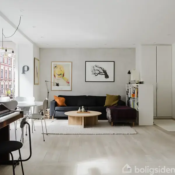 Pianolænestol i moderne stue, placeret ved vindue. En sofa med puder og et rundt sofabord dominerer midten. Lys indretning med kunst på væggen og reol til højre.