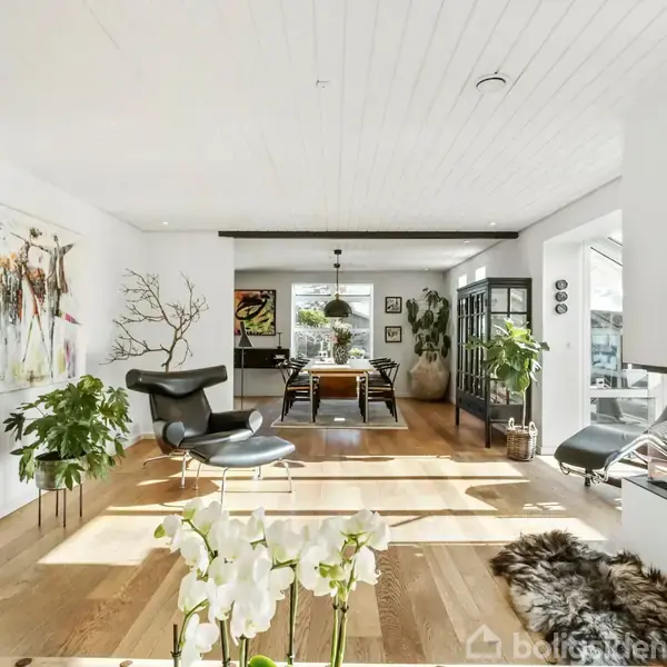Stue med moderne indretning, ild i pejsen, træmøbler og malerier på væggene. Store vinduer giver lys, og grønne planter skaber en naturlig atmosfære. En spiseplads er synlig i baggrunden.