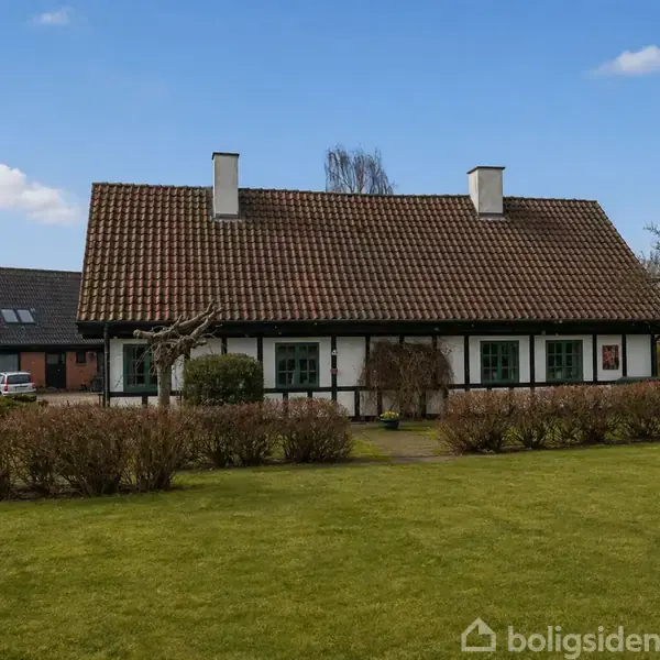 Et bindingsværkshus med to skorstene står i en velplejet have. Lav hæk omgiver huset i et landligt og roligt miljø under en klar blå himmel.