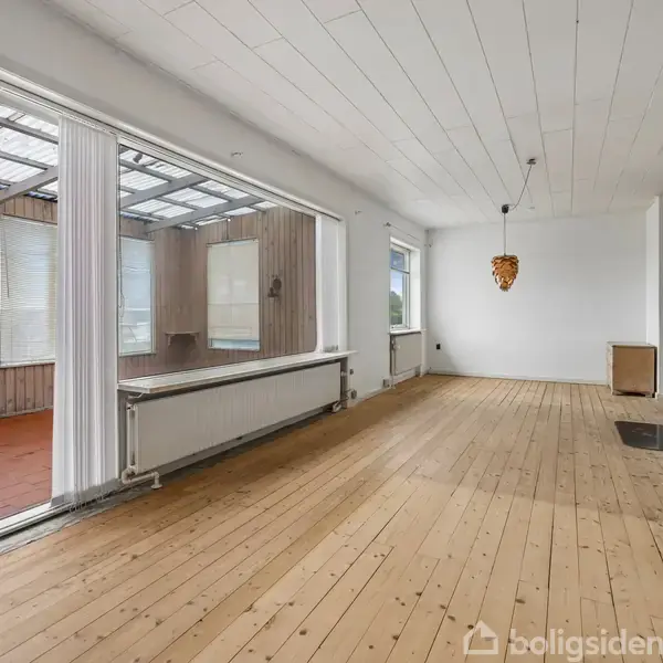 Stue med trægulv og brændeovn i et lyst rum. Til venstre er der et solarium med store vinduer og rødt gulv, hvilket giver naturligt lysindfald.