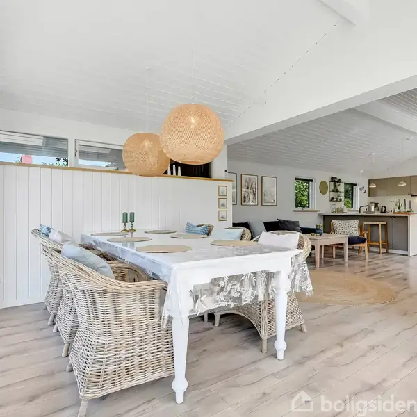 Et lyst rum med et spisebord dækket med dug og kurvestole. To runde lamper hænger over bordet. Køkkenet i baggrunden har moderne skabe og apparater.