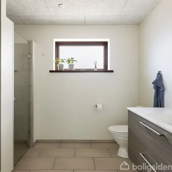 Badeværelse med moderne vaskbord og skabsarrangement til højre, toilet og bruser til venstre. Et lille vindue med planter over fliserne bringer lys ind.