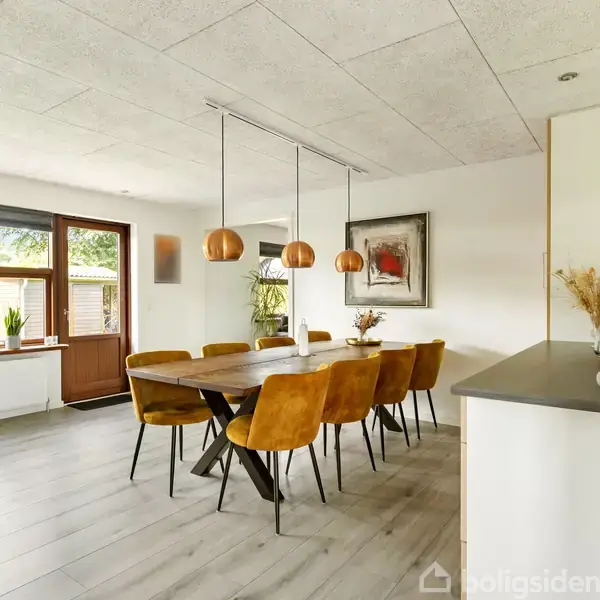Træbord omgivet af otte gule stole står i moderne spisestue. Tre kobberlamper hænger over bordet. Rummet har kunst på væggen og store vinduer med udsigt til haven.