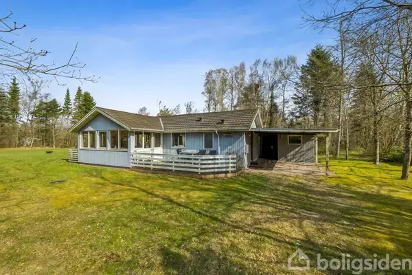 Et lyst, enkelt hus med terrasse står stille omgivet af en græsplæne og træer under en blå himmel.